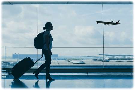 The silhouette of a female passenger pulling a luggage cart in an airport terminal with a flying plane in the background.